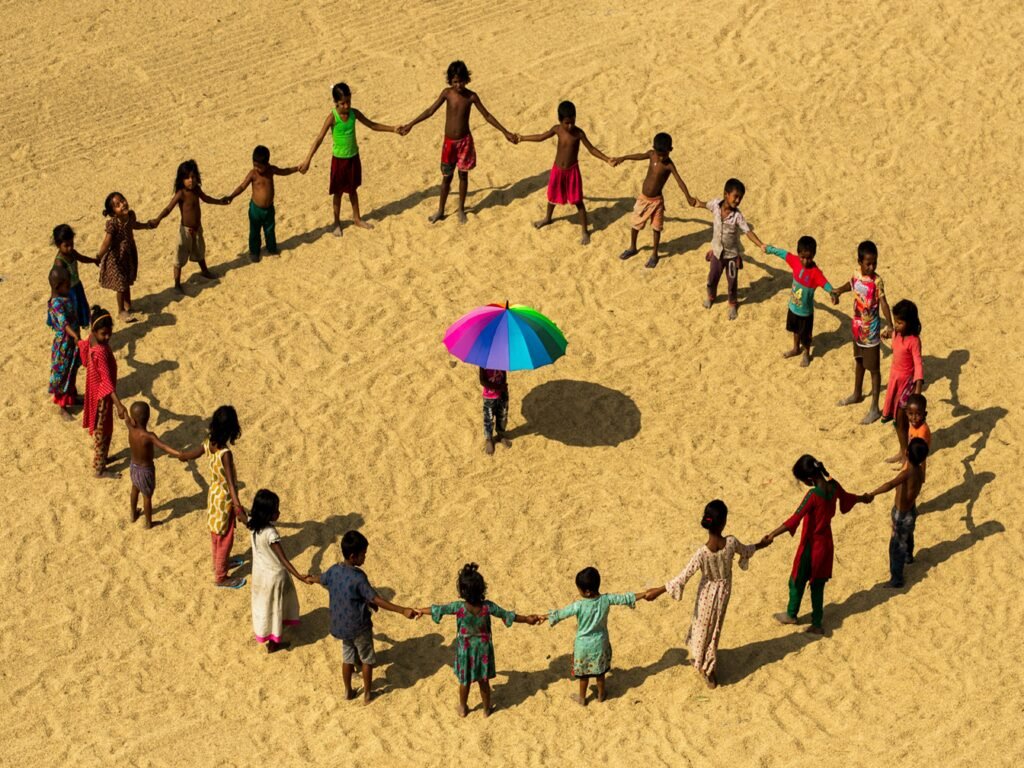 Aerial view of children holding hands in a circle on sandy ground with colorful umbrella in Chittagong, Bangladesh.